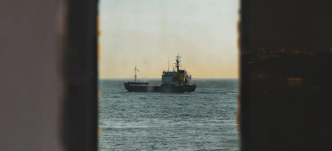 A small ship on calm water viewed through a rectangular opening, with soft sunset light on the horizon framed by dark edges.