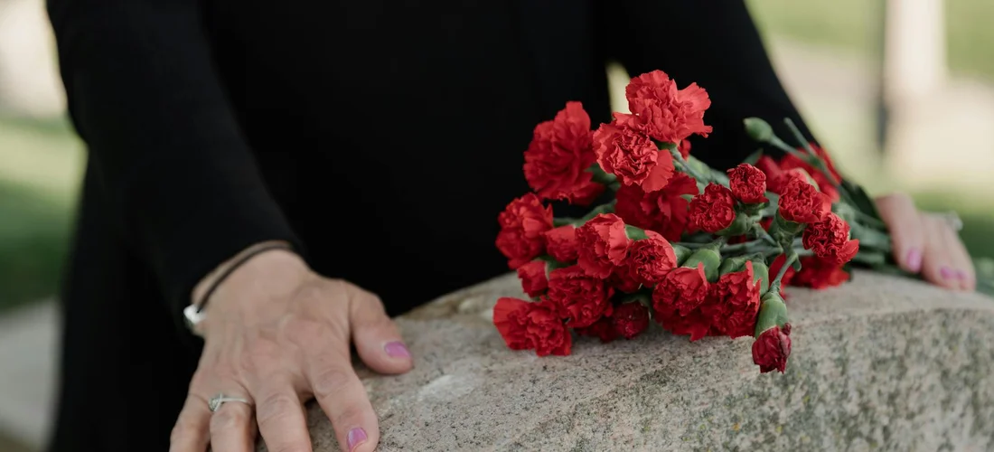 Close-up of a mourner in black clothing resting a hand on a stone ledge with a bouquet of red carnations placed on it, symbolizing mourning in Sikh funeral practice.