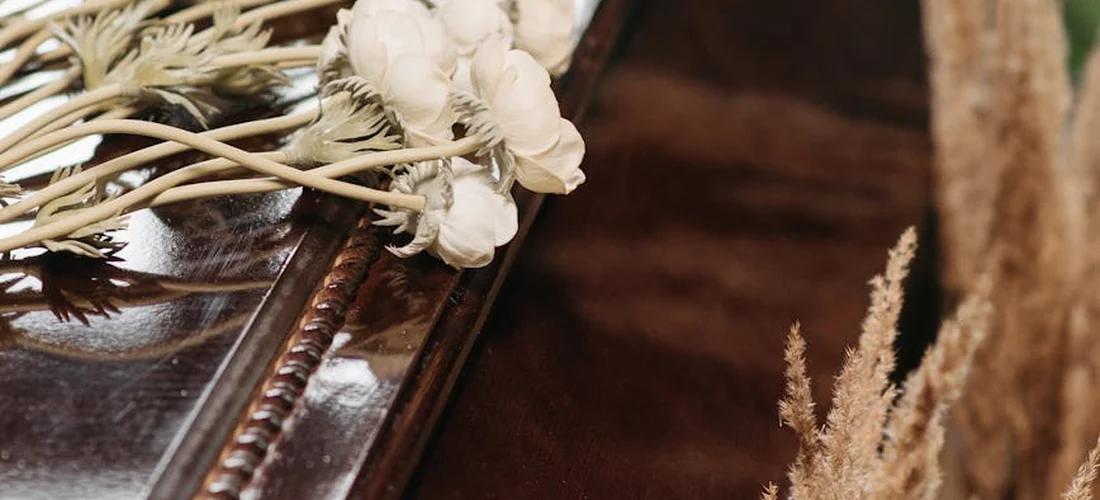 Close-up of a simple white floral casket spray resting on a polished wooden casket, accented with dried beige grasses.