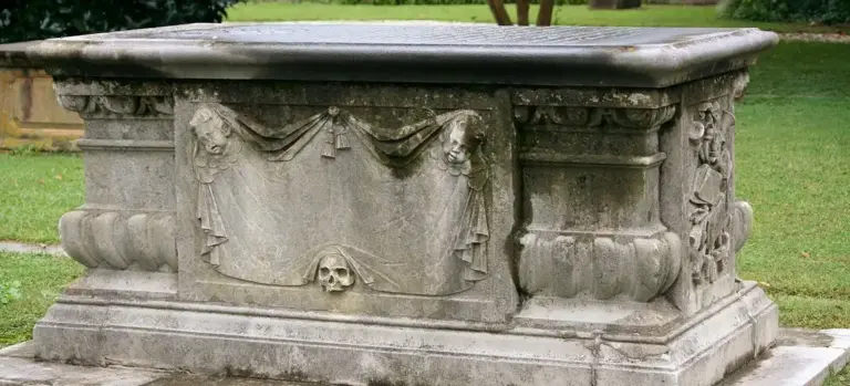 Carved stone funeral sarcophagus in a cemetery with draped decorations and a skull motif.