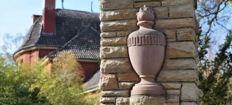 Stone urn mounted on a brick pillar with a house and greenery in the background.