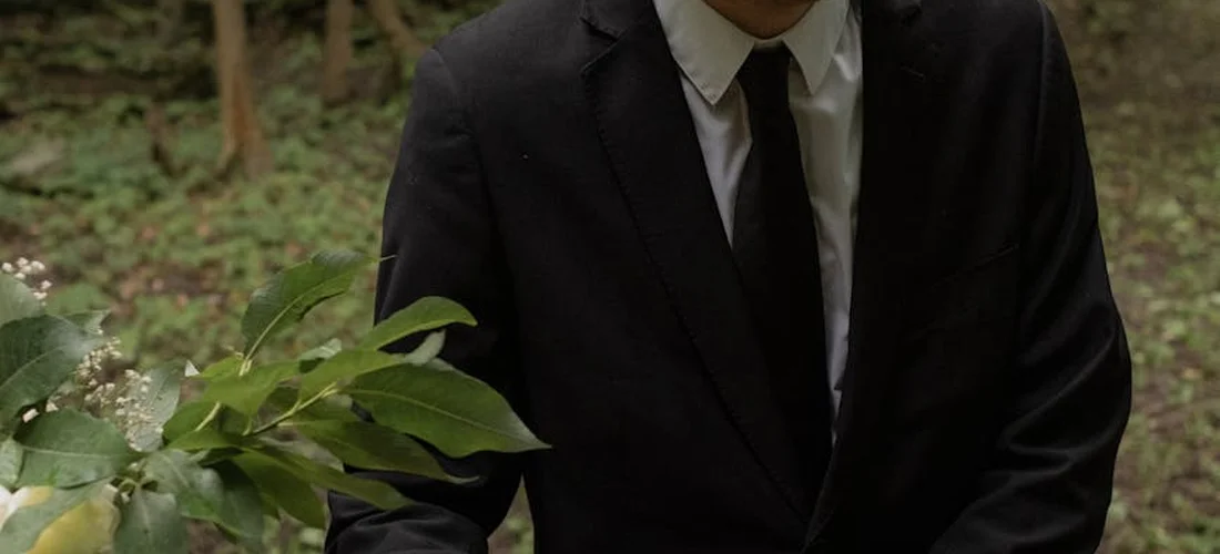 A person wearing a black suit and tie stands outdoors in a wooded area with greenery.