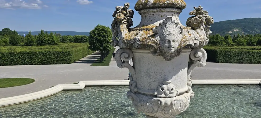 Ornate stone urn with carved faces atop a fountain in a formal garden, surrounded by hedges and a shallow pool