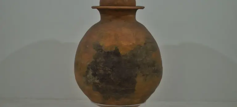 Brown ceramic cremation urn with dark patina marks on a neutral background; simple, rounded body and narrow neck.