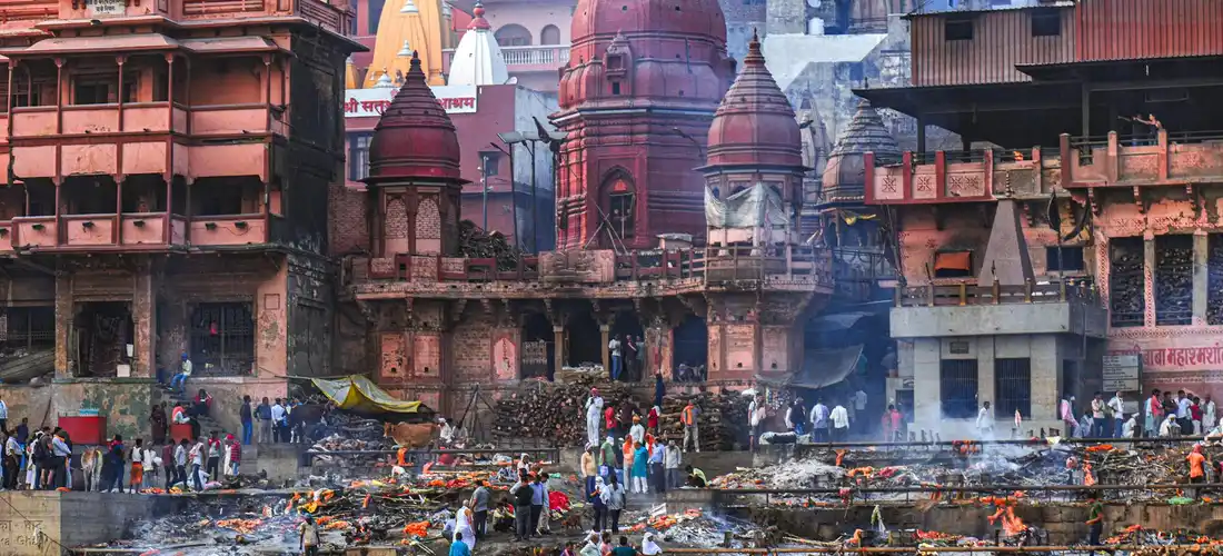 Cremation ghats along the Ganges River in Varanasi with burning pyres, scaffolding, and crowds.