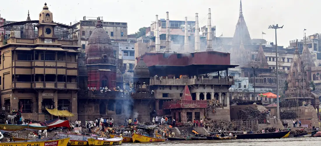 Riverside cremation ghats on the Ganges in Varanasi with several funeral pyres and smoke rising, crowded by boats and onlookers.