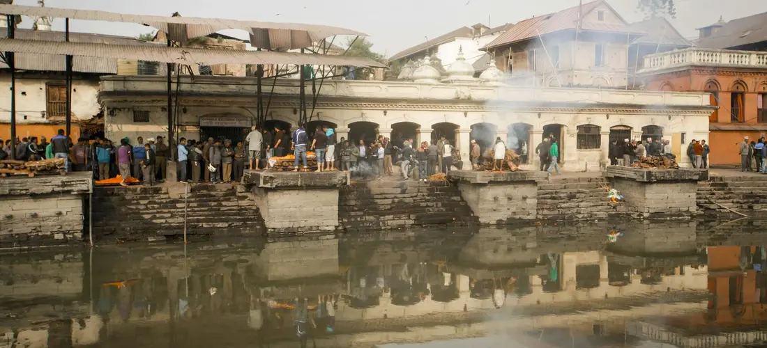 People gather along the riverfront cremation ghats with smoke rising and buildings reflected in the water.