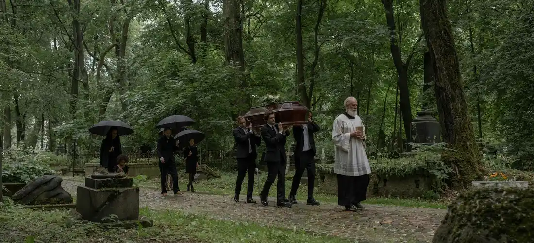 Funeral procession in a cemetery with pallbearers carrying a casket under umbrellas and a clergy member leading the service