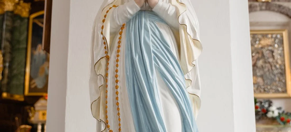 Statue of the Virgin Mary inside a church, wearing a blue robe and white mantle, with religious artwork in the background