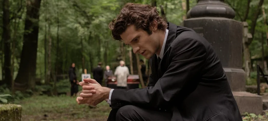 Man in a dark suit crouches in a cemetery, checking his wristwatch during a quiet memorial moment.