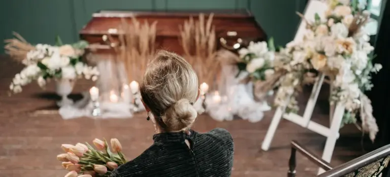 From behind view of an older person with gray hair wearing a dark sweater, looking toward a casket in a funeral home with floral arrangements.