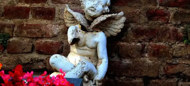 White winged cherub statue seated against a brick wall with red flowers in the foreground.