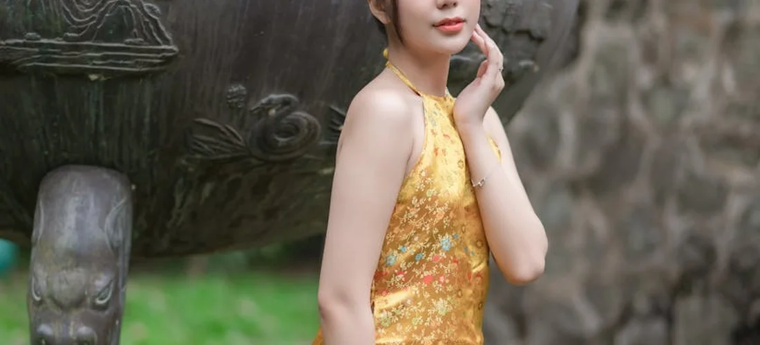 A person wearing a yellow floral dress stands beside a large stone urn in a garden.