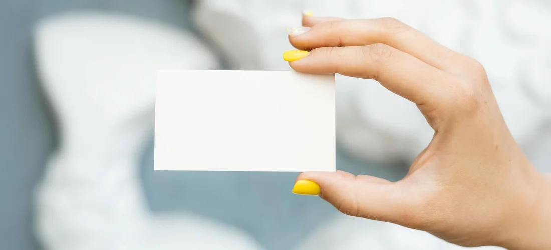 Close-up of a hand with yellow nails holding a blank white card, suggesting gathering essential information for memorial cards.