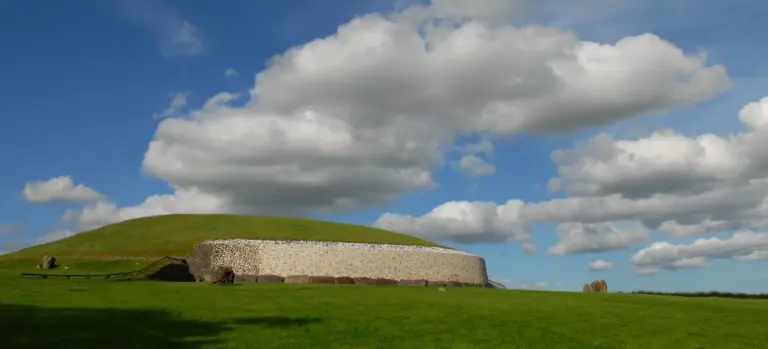 Grass-covered burial mound with a stone retaining wall along its side, under a bright blue sky with scattered clouds.