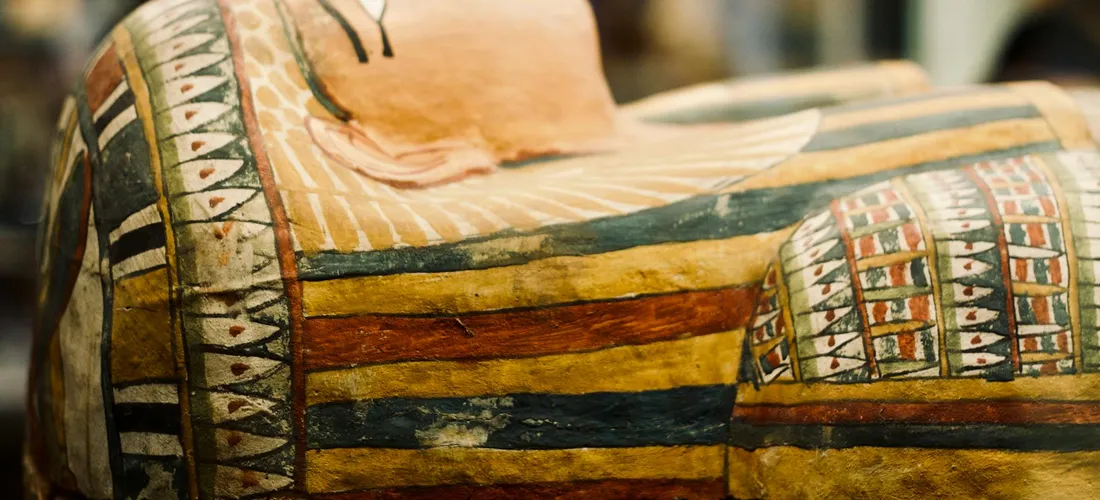 Close-up of a fabric burial shroud with horizontal stripes and geometric motifs in warm earth tones of ochre, blue, orange, and black.