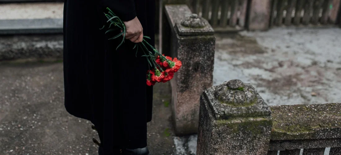 A person in dark clothing holding red flowers beside weathered gravestones in a cemetery.
