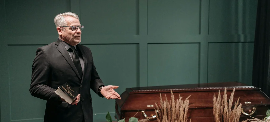 A clergyman in a black suit stands beside a closed wooden casket during a funeral service, holding a Bible and addressing attendees.
