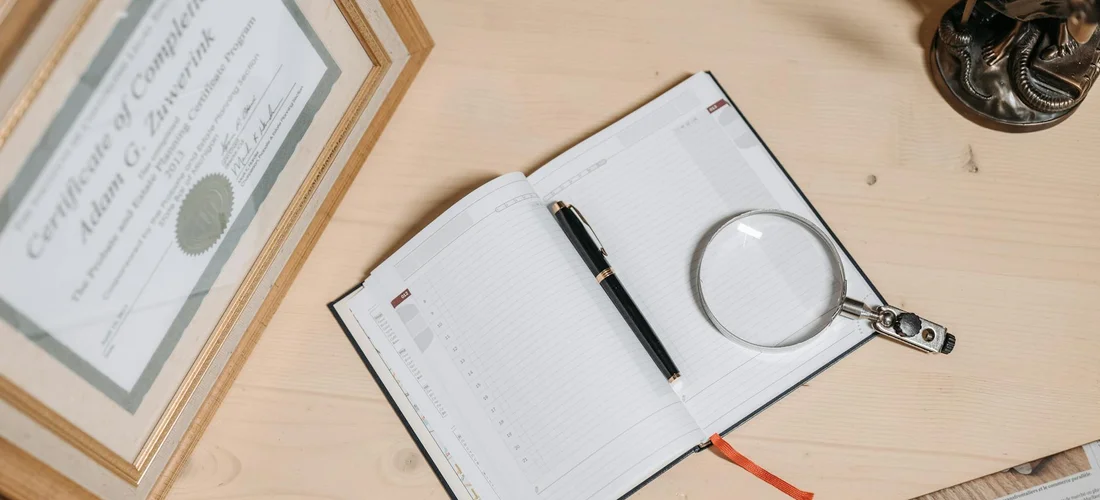Framed cremation certificate on a wooden desk beside an open notebook, a magnifying glass, and a pen.