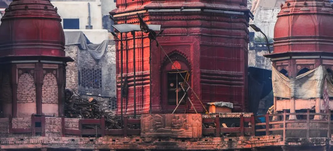 Cremation pyres along a riverbank at a Hindu cremation site, framed by red brick structures and wooden platforms.