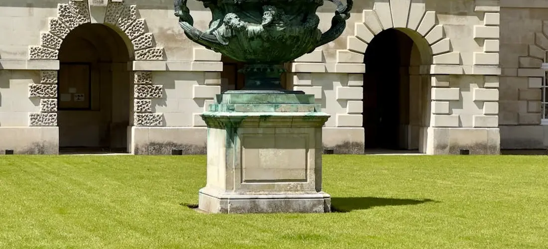Bronze urn on a pedestal in a stone courtyard with arched doorways and manicured lawn