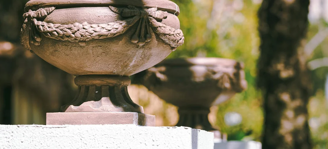 Outdoor urns displayed on a stone pedestal, bathed in warm sunlight among greenery.