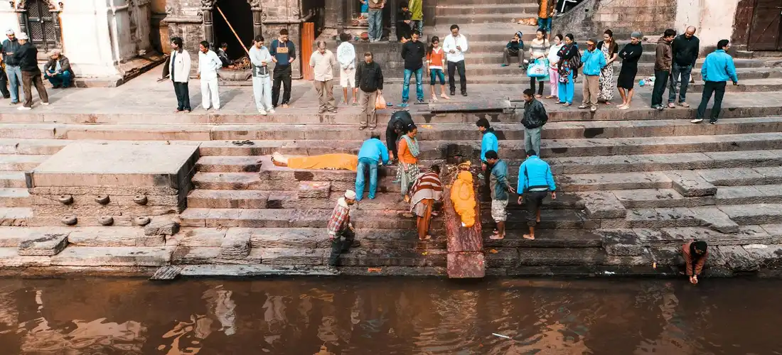 A diverse group of people stands along stone steps by a river, participating in or observing a cremation rite.