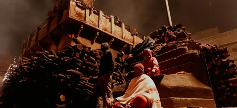 Mourners gathered near a cremation site with a wooden platform and stacked logs, as a subdued, smoky atmosphere surrounds the ceremony.