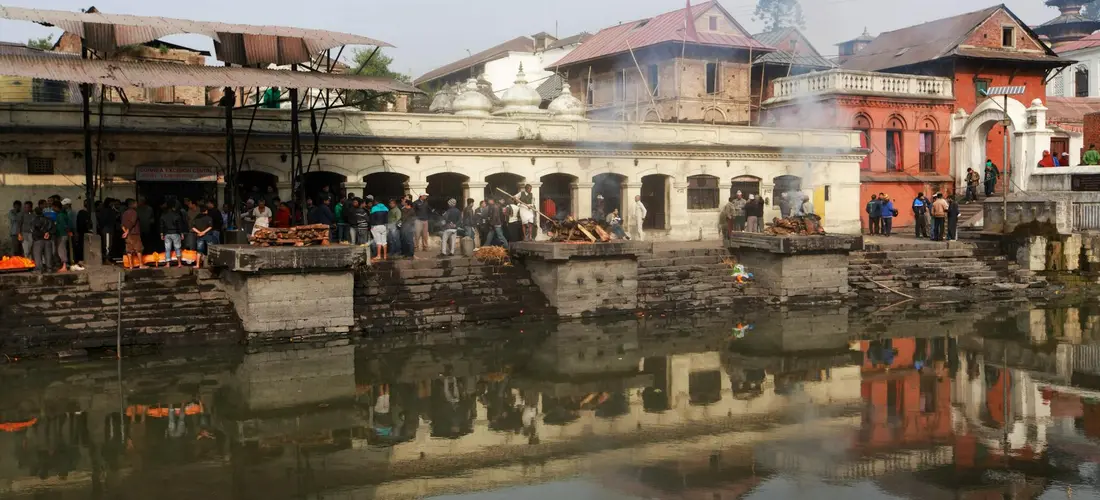 People gathered on stone steps along a river at a cremation ghat with platforms and temple buildings in the background.