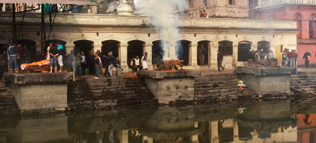Cremation pyres along a riverbank with smoke rising, spectators nearby.