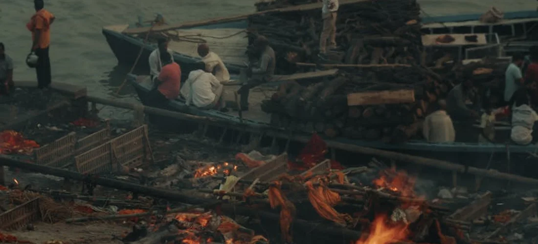 Riverside cremation scene with people standing near burning funeral pyres and boats docked in the background.