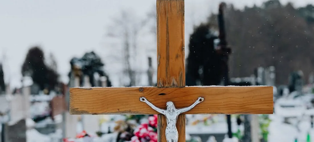 Close-up of a wooden cross with a metal figure of Jesus in a snow-covered cemetery with gravestones in the background.