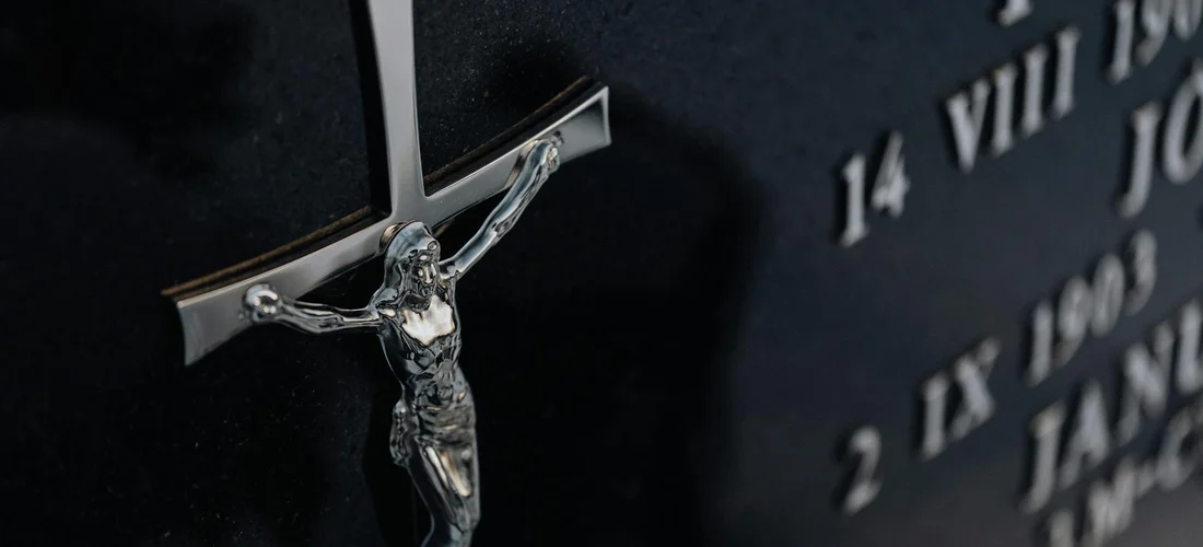 Close-up of a metallic crucifix mounted on a dark memorial stone with engraved numbers blurred in the background.