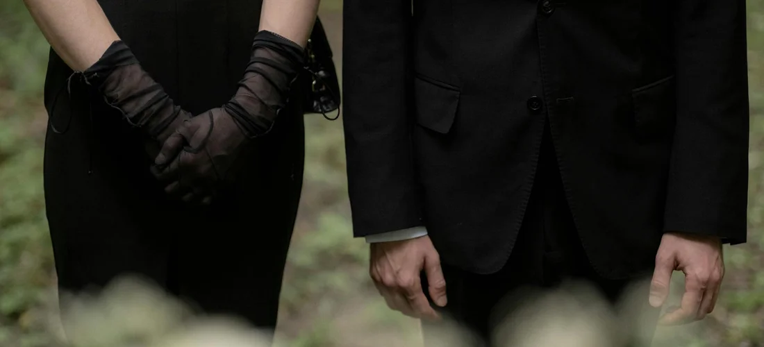 Two people in black formal attire standing with gloved hands, suggesting a funeral or memorial scene.