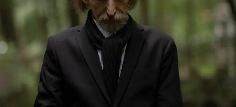 A person wearing a black suit and scarf stands in a blurred outdoor setting, representing mourners and considerations of funeral attendance and privacy.