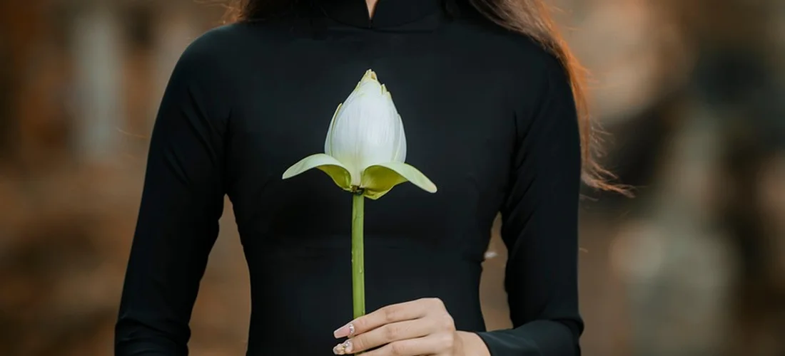 Person wearing a black modest outfit holding a white calla lily against a blurred background.