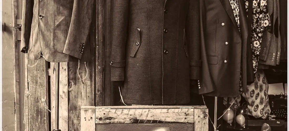 Sepia-toned display of formal coats and suits hanging on racks in a rustic shop.