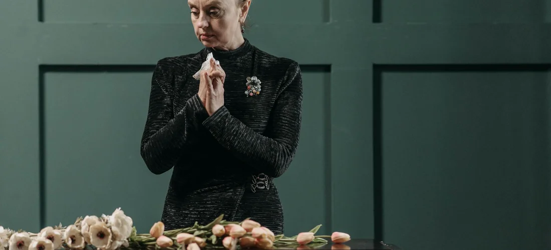 Person in dark clothing stands with hands clasped in front of a table of pale flowers, against a green paneled wall.