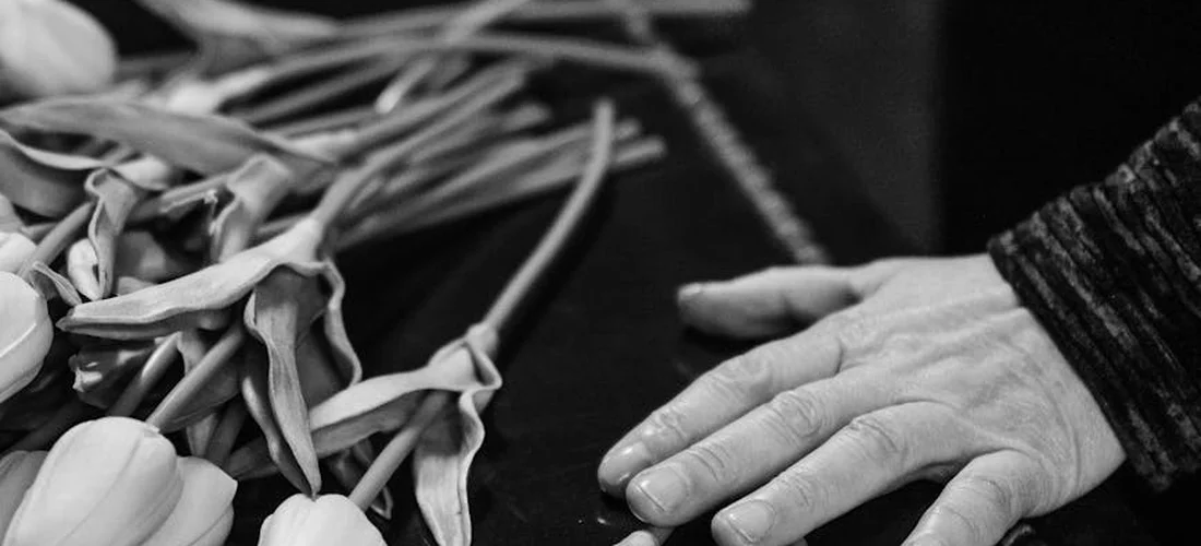 Close-up of a hand resting beside a bouquet of flowers in black and white, conveying a funeral setting.