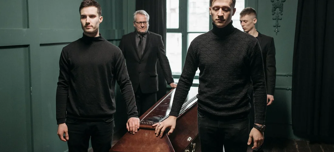 Four pallbearers in black clothing stand around a closed coffin in a dimly lit funeral home.