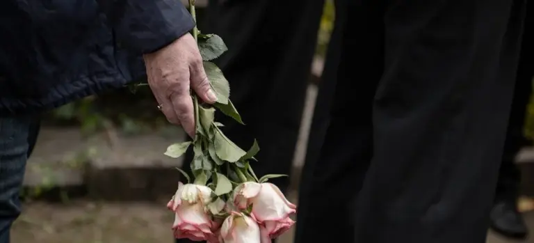 Close-up of a hand holding a bouquet of pale pink and white roses at a funeral, with mourners in dark clothing in the background.