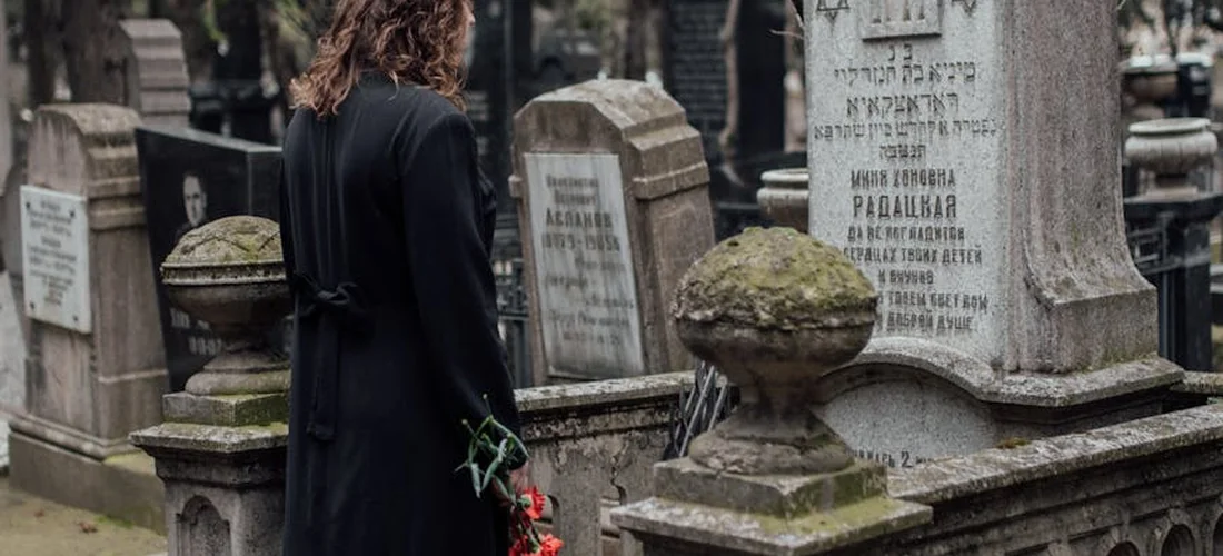 A person in a dark coat stands by weathered gravestones in a cemetery, holding a small bouquet of red flowers.