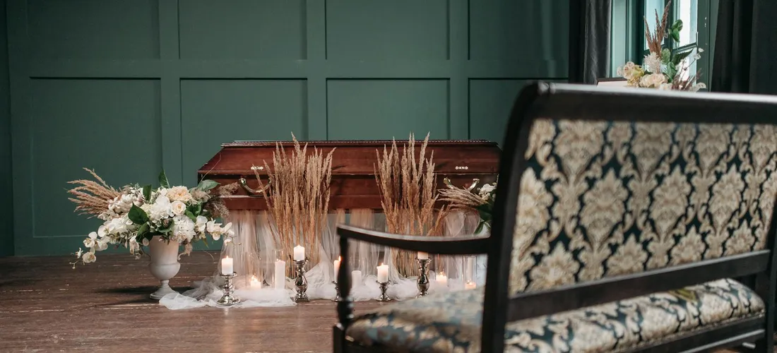 Casket in a funeral room with white floral arrangements, tall candles, and a patterned chair in the foreground.
