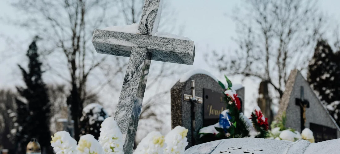 Grave markers in a cemetery including a prominent cross-shaped tombstone surrounded by flowers