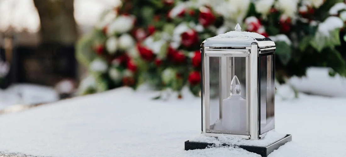 Lantern resting on a snow-covered surface with blurred red and white funeral flowers in the background.