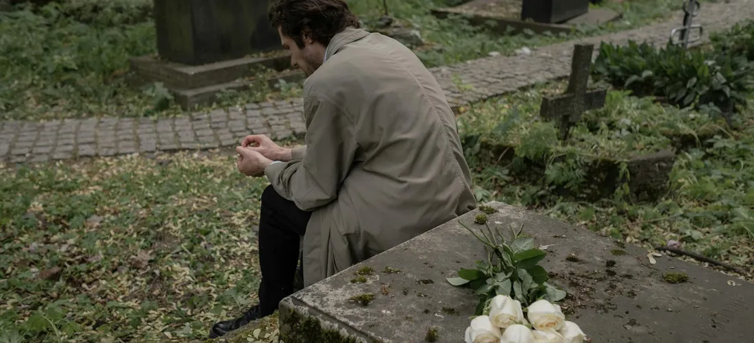 A person sits on a stone bench beside a gravestone in a cemetery, head bowed in contemplation, with flowers resting on the stone.