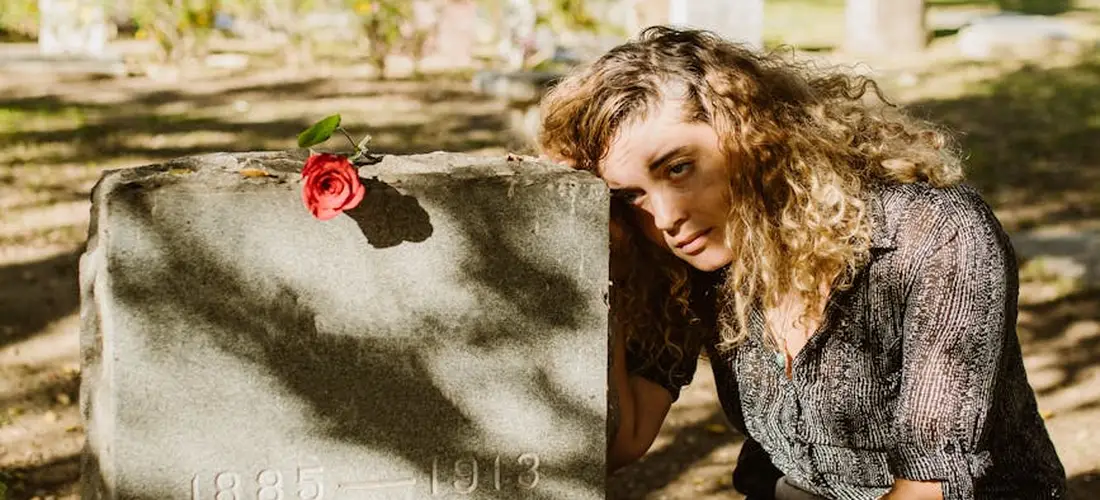 A woman with curly hair sits beside a gravestone in a cemetery, a single red rose resting on the stone, conveying a somber, reflective mood.