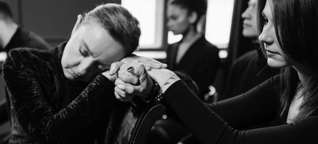 People at a funeral hold hands in a quiet, intimate moment, suggesting a private, respectful setting.