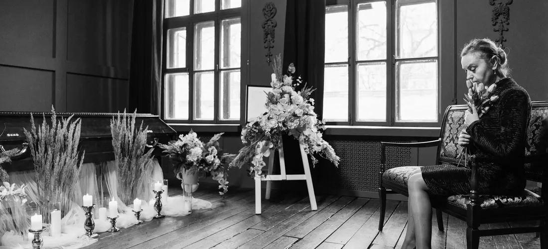 A subdued funeral scene with a person seated in a chair, wearing dark sunglasses, surrounded by flowers and candles.