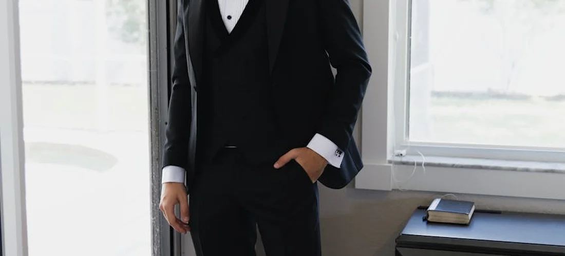 Man in a black suit with a white dress shirt and black vest, standing indoors near a window, illustrating a formal, coordinated look for a funeral.